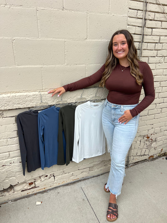 Woman standing next to a wall with hanging clothes, wearing a brown long-sleeve top and light blue jeans.