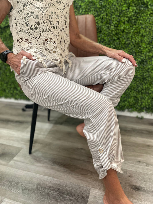 Person wearing a white lace top and striped pants sitting on a chair with a green bush background.