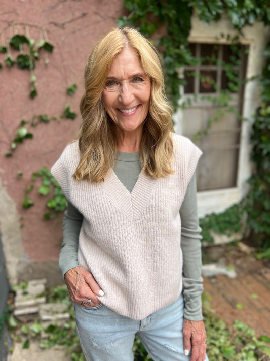 Woman wearing a light pink vest over a gray long-sleeve shirt, standing outdoors with greenery in the background.